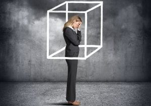 A businesswoman stands with her head in her hands as she is surrounded by a mixed media cube.  She is feeling as though she is boxed in by matters beyond her control.